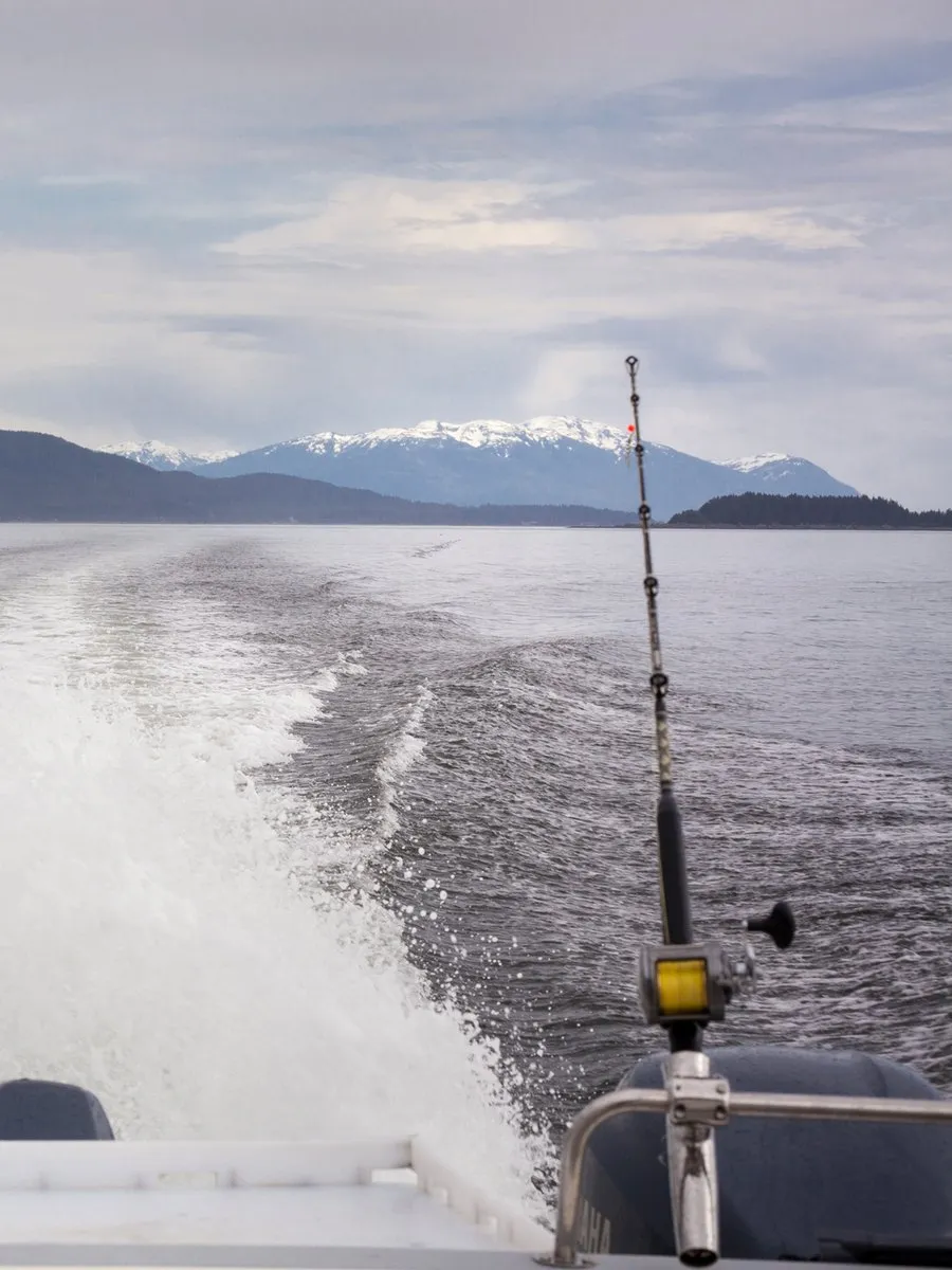 A heavy rod upright in a stern rod holder, the boat's wake running back towards a snow-covered ridge